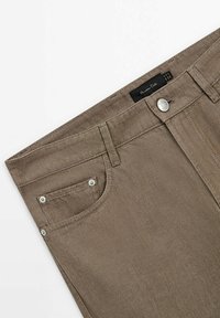 Jean en denim marron montrant la partie avant supérieure avec la ceinture, le bouton, les passants de ceinture, une petite poche avant et une étiquette de marque visible à l'intérieur de la ceinture.