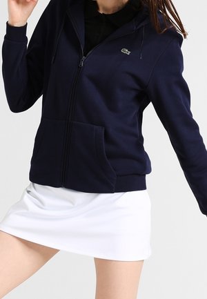 Persoon draagt een marineblauwe hoodie met rits en een logo, gecombineerd met een witte rok, poseert met één arm omhoog tegen een effen achtergrond.