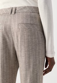 Close-up of a person wearing beige pinstriped trousers and a long-sleeve cream top, showing back pockets and right hand beside the hip.