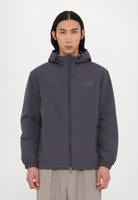 Mørkegrå zip-up jakke med hætte, elastiske manchetter og et logo-patch på brystet. Glat mat tekstur, afslappet pasform og to sidelommer.