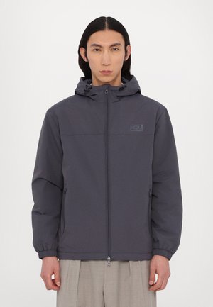 Mørkegrå zip-up jakke med hætte, elastiske manchetter og et logo-patch på brystet. Glat mat tekstur, afslappet pasform og to sidelommer.