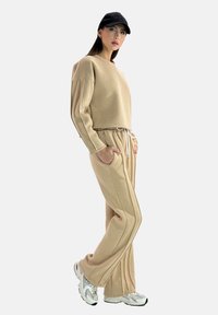 Femme portant un ensemble de vêtements d'intérieur beige avec des manches longues et un pantalon à cordon, des baskets blanches et une casquette de baseball noire, debout les mains dans les poches.