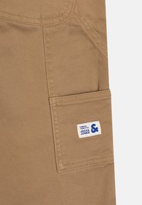 Beige stoff med en glatt tekstur, med en stor sidelomme og en blå-hvitt merket etikett "JACK & JONES" sydd på.