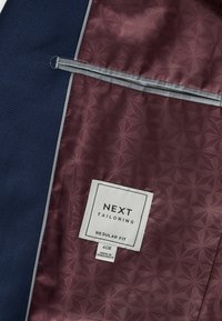 Tumes tekstuuriga kangast jope sisemus burgundi mustriga voodriga, halli ääristuse ja valge sildiga, millel on kirjas "NEXT TAILORING REGULAR FIT."