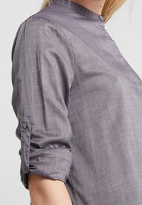 Košile s button-up zapínáním z texturované světle šedé tkaniny, s vyvýšeným límcem a ohrnutými rukávy zajištěnými knoflíky.