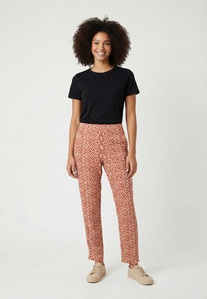 Femme souriante aux cheveux bouclés portant un t-shirt noir à manches courtes, un pantalon rouge et blanc à motifs, et des baskets beiges, debout devant un fond uni.
