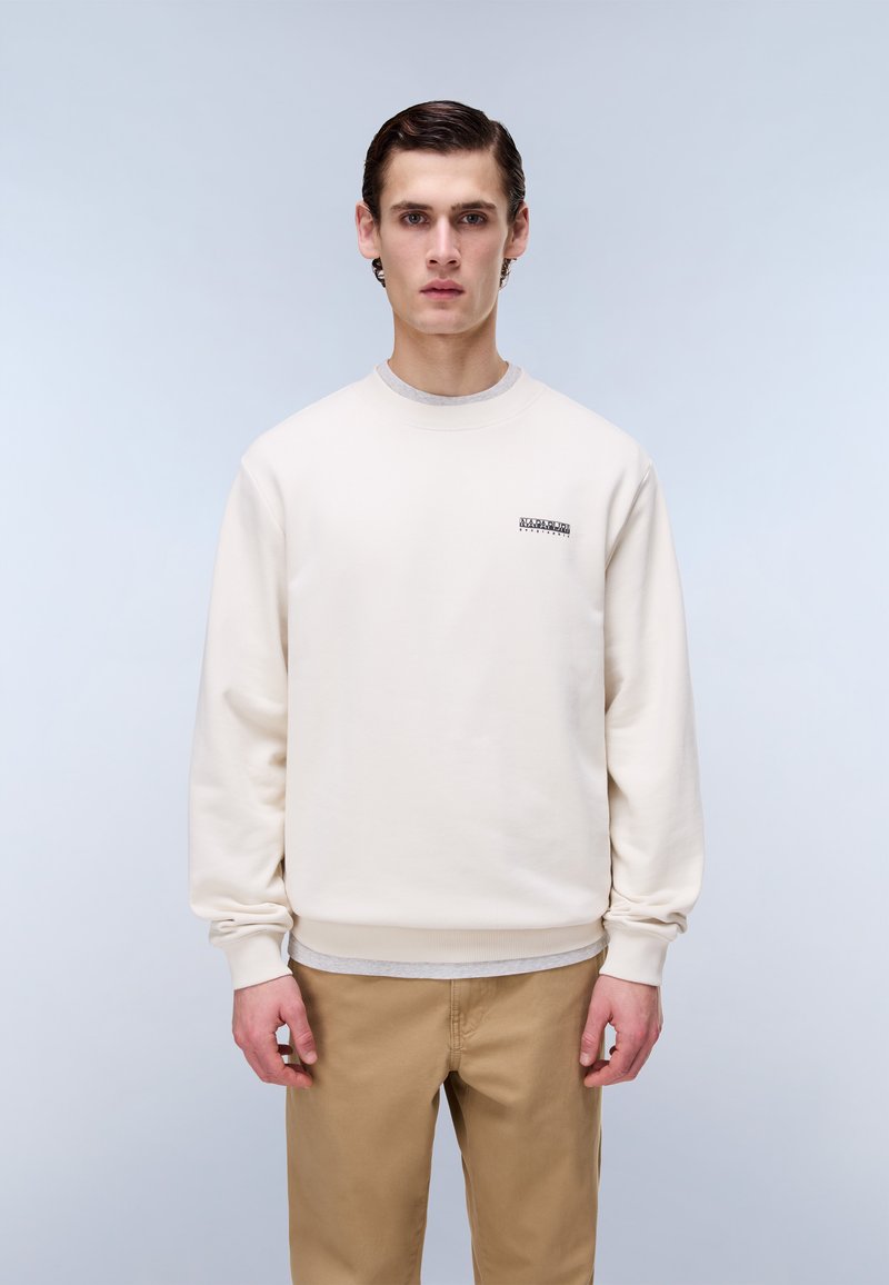 Junger Mann steht still, trägt einen cremefarbenen Pullover und hellbraune Hosen vor einem schlichten, hellgrauen Hintergrund.