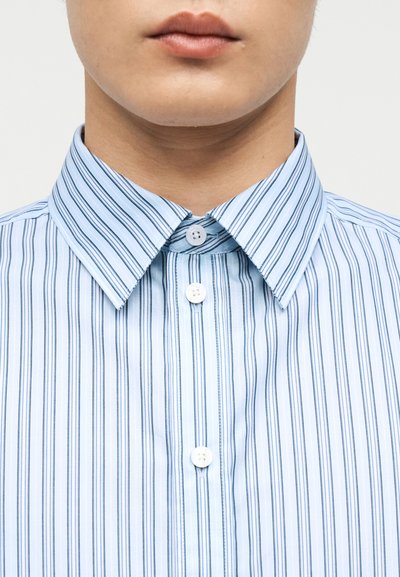 Κοντινό πλάνο του κάτω μέρους του προσώπου και του λαιμού ενός ατόμου που φοράει ανοιχτό μπλε πουκάμισο με σκούρες κάθετες ρίγες και λευκά κουμπιά.