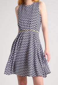Robe à motif chevron marine et blanche avec un design sans manches, taille ajustée et jupe évasée. Présente un accent jaune à la taille.