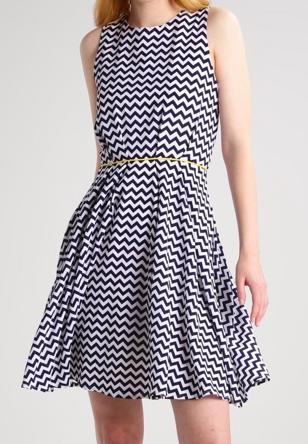 Robe à motif chevron marine et blanche avec un design sans manches, taille ajustée et jupe évasée. Présente un accent jaune à la taille.