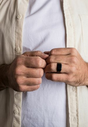 Anello rettangolare nero sulla mano, mani unite. Camicia chiara sopra una maglietta bianca, mostrando texture e colori.