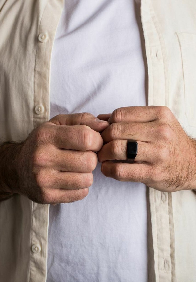 Schwarzer rechteckiger Ring an der Hand, Hände zusammengelegt. Hellfarbige Hemdbluse über einem weißen T-Shirt, das Texturen und Farben zeigt.