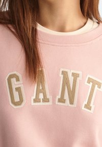 Sweatshirt rose clair avec de grandes lettres brodées beiges formant le mot "GANT". Le tissu est doux avec des détails de col côtelé.