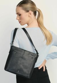 Czarna skórzana torebka o teksturowanym wykończeniu, z długim paskiem i tylną kieszenią, noszona przez osobę w jasnoniebieskim swetrze.