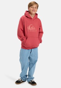 Rood hoodie met een groot logo op de voorkant, gecombineerd met lichtblauwe, losse jeans en zwarte sneakers met witte accenten.