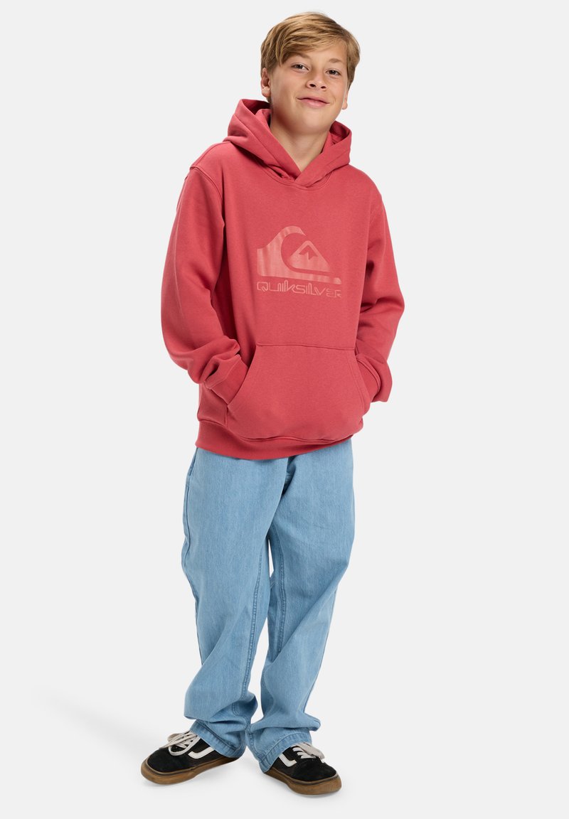 Rood hoodie met een groot logo op de voorkant, gecombineerd met lichtblauwe, losse jeans en zwarte sneakers met witte accenten.
