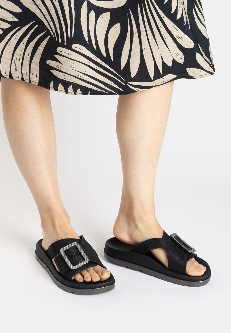 Black slide sandals made of synthetic material featuring a large metallic buckle, soft footbed, and textured straps, worn with a patterned skirt.