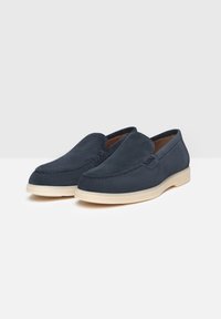 Zapatos negros de ante sin cordones con puntera redondeada, costuras decorativas y una suela de goma beige. Textura suave, diseño minimalista.