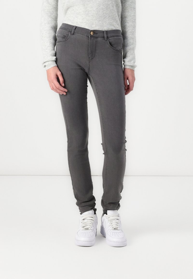 Jean skinny gris en tissu extensible avec cinq poches et fermeture à bouton à l'avant, associé à des baskets blanches.