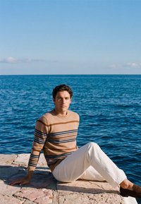 Jeune homme portant un pull rayé et un pantalon blanc assis sur un pavé en pierre au bord d’une mer bleue calme sous un ciel clair.