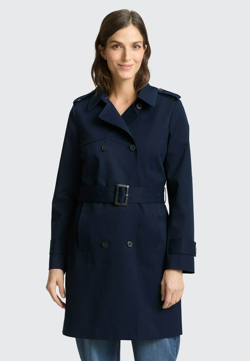 Frau trägt einen marineblauen, doppelt geknöpften Trenchcoat mit Gürtel, steht vor einem einfachen hellen Hintergrund und lächelt leicht.