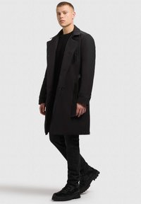 Juodas paltas su struktūrizuota dizainu, pasižymintis blizgiu apykakle ir šoninėmis detalėmis. Dėvimas ant juodos megztinio ir džinsų, derinamas su masyviomis batais.