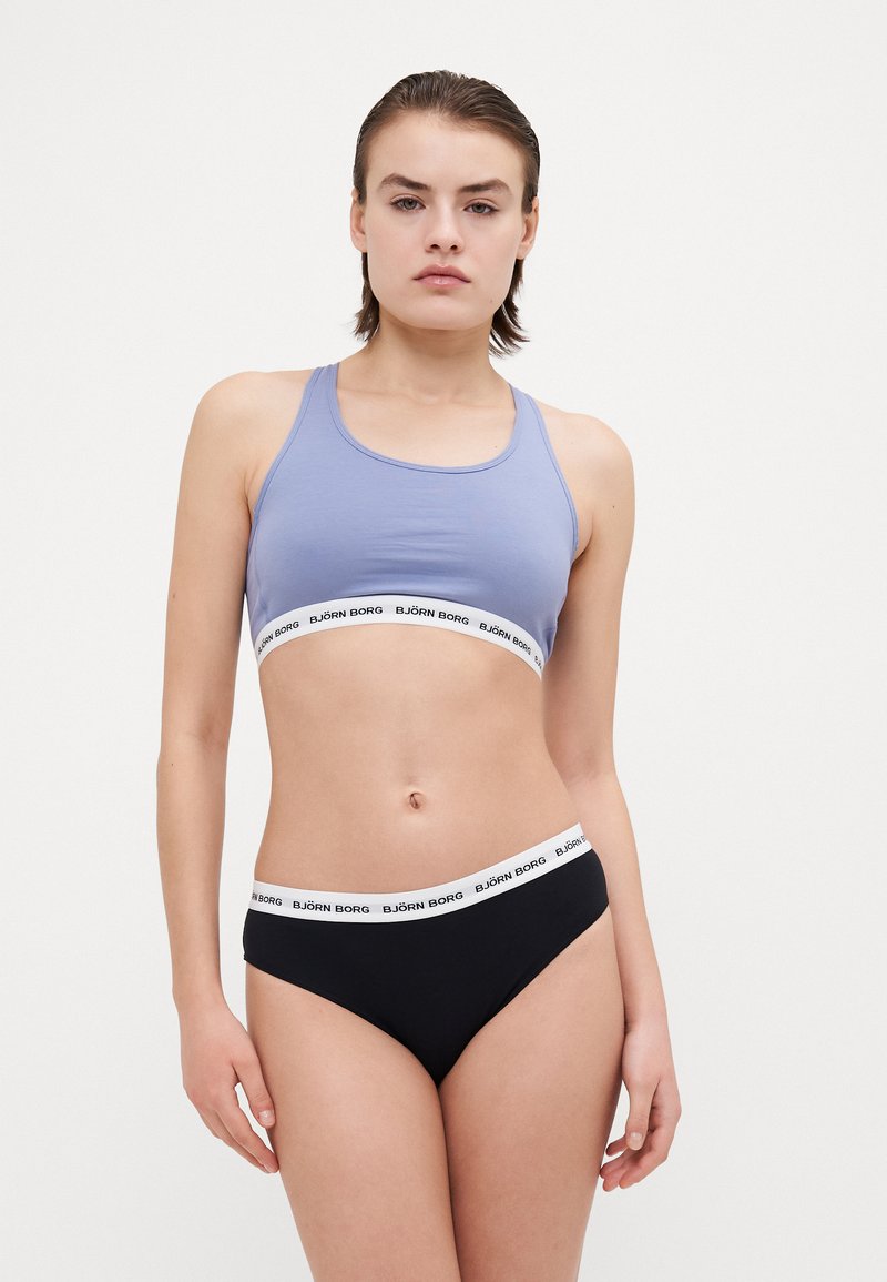 Helles blaues Sport-BH mit einem weißen Markenband; kombiniert mit schwarzen Slips, die ein passendes weißes Band haben. Glattes, dehnbares Material.