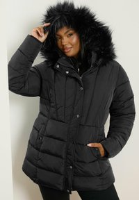 Svart puffjacka med en fuskpäls huva, quiltad textur, knapp- och dragkedjestängning samt två sidofickor.