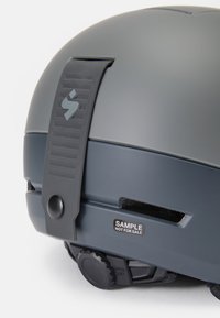 Casco en gris oscuro mate con una correa de ajuste texturizada en negro. Cuenta con un dial para el tamaño y una etiqueta que dice "MUESTRA NO A LA VENTA."
