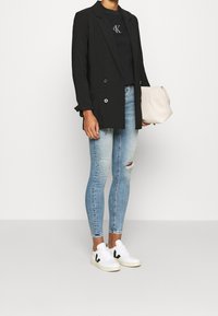 Schwarzer zweireihiger Blazer über einem schwarzen T-Shirt, kombiniert mit hellblauen, zerrissenen Skinny-Jeans und weißen Sneakers mit schwarzen Akzenten. Hält eine beiger Tasche.