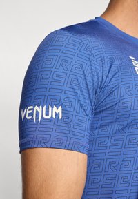 Μπλε αθλητικό πουκάμισο με γεωμετρικό σχέδιο. Διαθέτει λευκό λογότυπο «VENUM» στο μανίκι. Κατασκευασμένο από λείο, ελαφρύ υλικό.