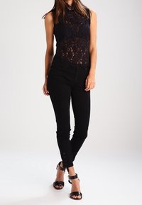 Svart spets utan ärmar med hög krage, ihop med åtsittande svarta byxor med knapppdetaljer vid anklarna och svarta klacksandaler.