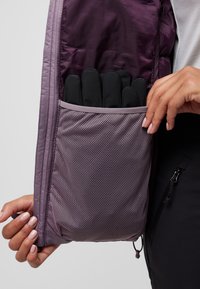 Interno di giacca viola con una tasca in rete contenente guanti neri. La tasca ha un bordo di un viola chiaro e il tessuto è liscio e trapuntato.