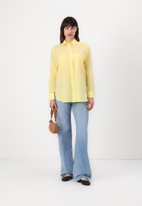 Camicia gialla con bottoni e tasca, indossata con jeans svasati azzurri chiari e ballerine nere, mentre tiene una piccola borsa marrone.