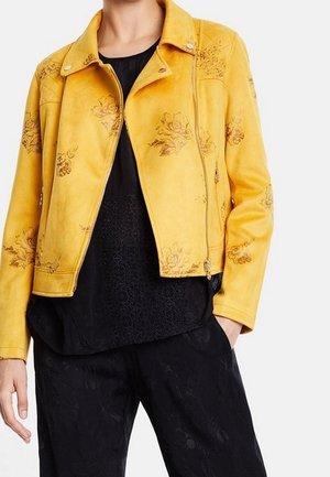 Personne portant une veste en suède jaune à motifs floraux sur un haut texturé noir et un pantalon noir, une main dans la poche.