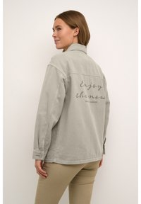 Grijze denimjas met lange mouwen, een kraag en de opdruk "Geniet van het nu" op de achterkant. Glad oppervlak en relaxte pasvorm.