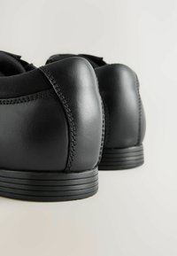 Baskets en cuir noir avec une texture lisse, talon arrondi et détails cousus. Semelle en caoutchouc avec des rainures horizontales pour l'adhérence.