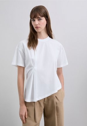 Camiseta blanca de manga corta con cuello redondo, que presenta un diseño drapeado en un lado, combinada con pantalones beige de talle alto.