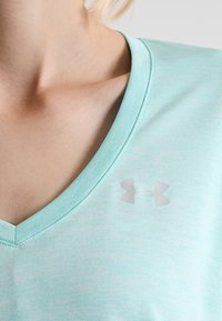 Närbild av en person som bär en ljusblå V-ringad tröja med en reflekterande Under Armour-logotyp på bröstet.