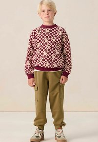 Un sweat-shirt bordeaux avec un visage souriant et un motif damier, associé à un pantalon cargo vert olive et des baskets beiges avec des accents multicolores.