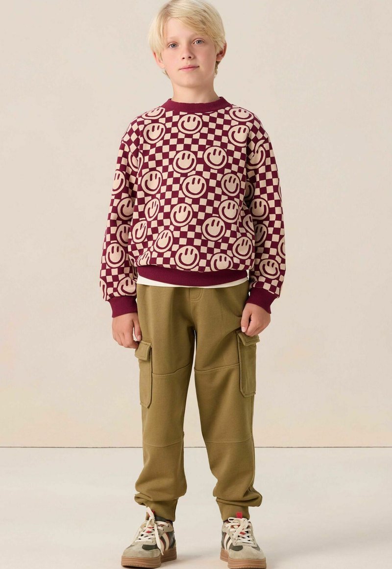 Un sweat-shirt bordeaux avec un visage souriant et un motif damier, associé à un pantalon cargo vert olive et des baskets beiges avec des accents multicolores.