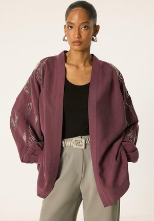 Femme portant un cardigan prune oversize avec des détails perlés sur les manches, un haut noir, un pantalon beige et une ceinture strass, debout devant un fond uni.