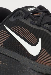 Black athletic shoe with textured knit upper, prominent white swoosh, brown accents, and a patterned rubber sole featuring speckled detail.
