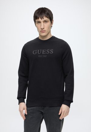 Młody mężczyzna ubrany w czarną koszulkę z długim rękawem z logo "GUESS EST. 1981", w zestawieniu z ciemnoszarymi dżinsami, stoi na tle jednolitej białej ściany.