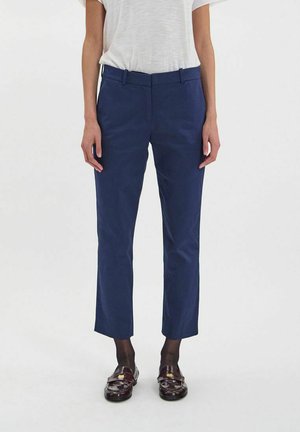 Femme portant un pantalon bleu marine longueur cheville, une chemise blanche à manches courtes, des chaussettes noires transparentes et des mocassins marron foncé, debout devant un fond uni.