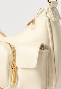 Sac à main en cuir crème avec des fermetures éclair couleur or et une poche avant cousue avec un rabat, présenté en gros plan.