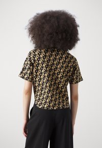 Personne aux cheveux bouclés portant un haut à manches courtes beige et noir à motifs et un pantalon noir, vue de dos sur un fond uni.