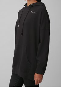 Hoodie noir en tissu doux, avec une poche avant, une capuche réglable à cordon de serrage et un petit logo sur la poitrine. Manches longues.