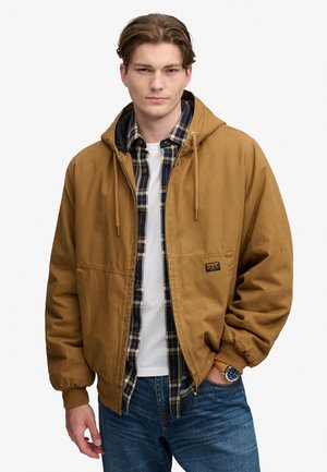 Ung mand iført en brun hættetrøje over en ternet skjorte og en hvid t-shirt, med blå jeans og et armbåndsur, stående mod en ensfarvet baggrund.