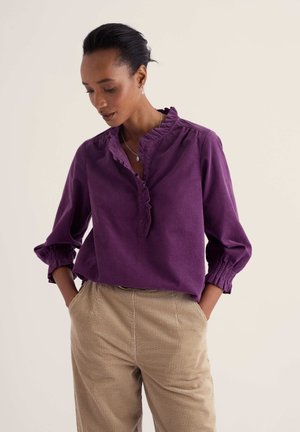 Femme portant un chemisier violet à volants avec des manches trois-quarts et un pantalon en velours côtelé beige, mains dans les poches, regardant vers le bas.